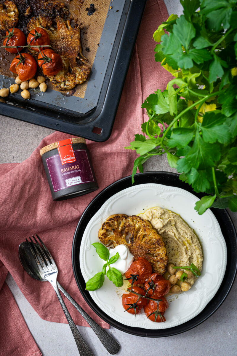 Gerösteter Blumenkohl mit gebackenen Tomaten, Kichererbsen und Hummus auf einem Teller, daneben frische Kräuter und Gewürz.