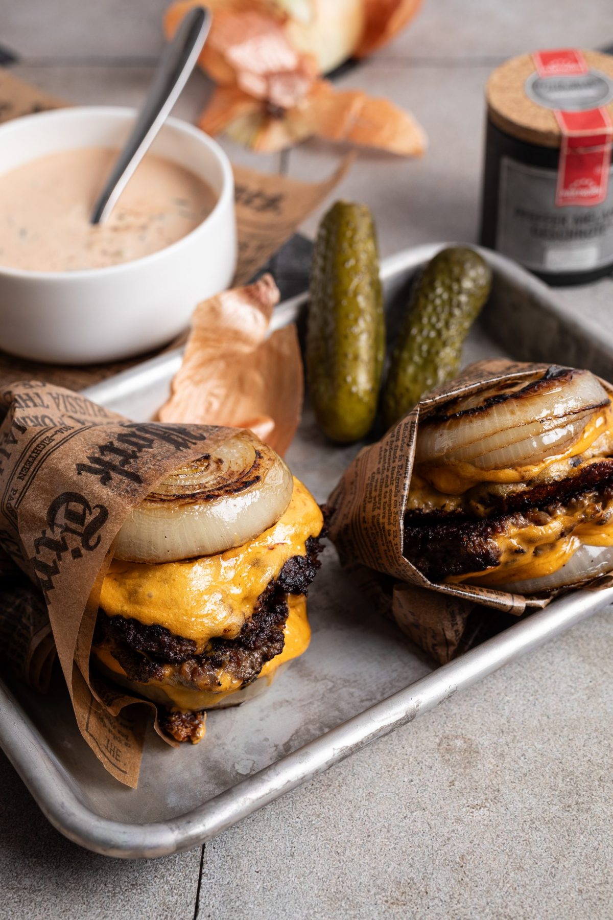 Zwei Cheeseburger mit gegrillter Zwiebel, in Papier gewickelt, auf Tablett mit Gurken und Schüssel mit Sauce daneben.