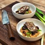 Zwei Schüsseln mit gefüllten Fleischrouladen auf Couscous, garniert mit Frühlingszwiebeln, neben einem Messer auf Holzbrett.