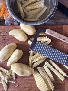 Wellenschneider und Rohkartoffeln auf Holzbrett, daneben geschnittene Kartoffeln und Topf mit Wasser.