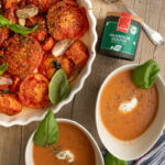 Zwei Schalen Tomatensuppe mit Sahne und Kräutern, daneben eine Auflaufform mit gebackenen Tomaten, Kräuterdose und Löffel.