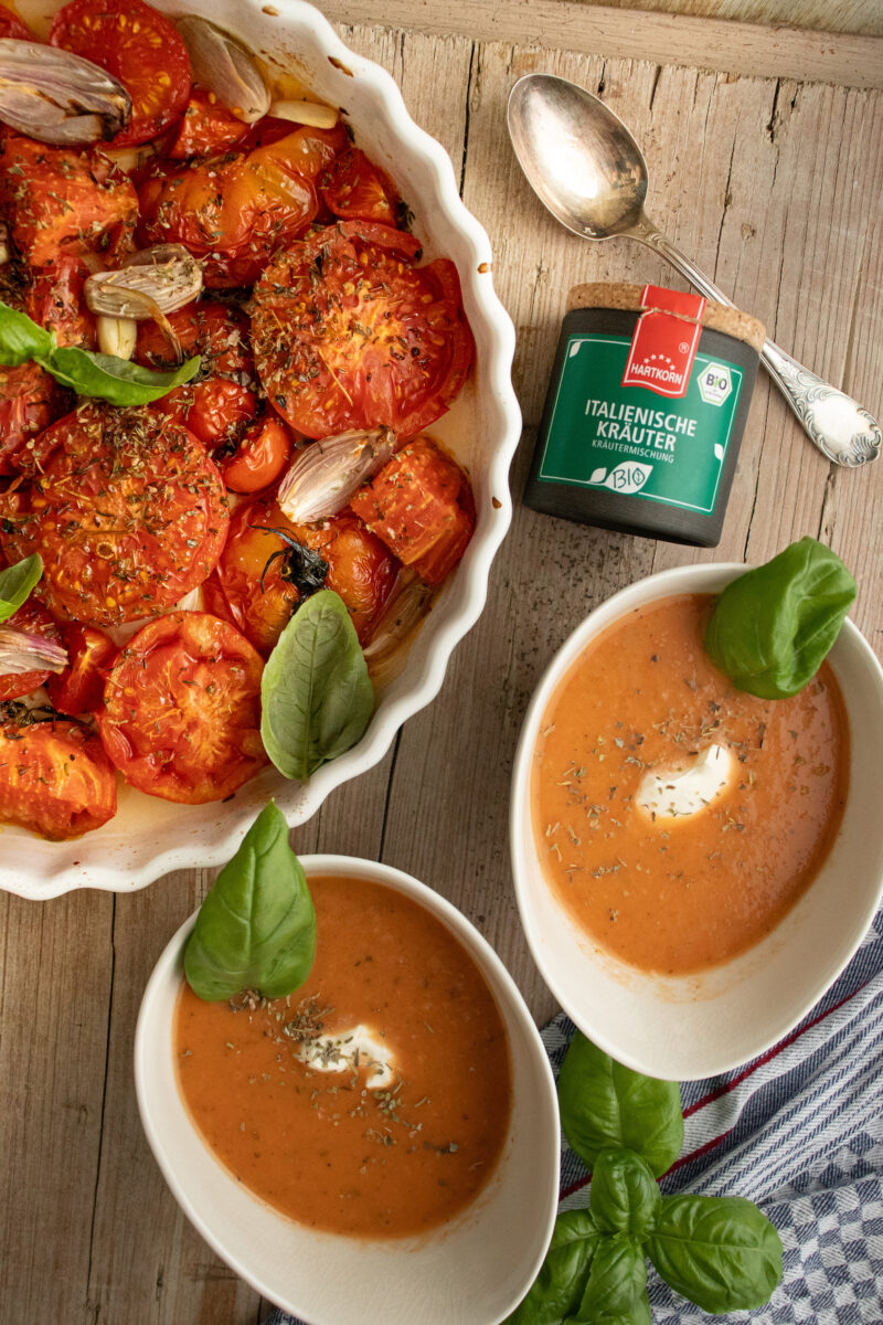 Zwei Schalen Tomatensuppe mit Sahne und Kräutern, daneben eine Auflaufform mit gebackenen Tomaten, Kräuterdose und Löffel.