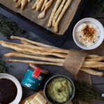 Knusprige Grissini auf Backblech, mit Dips, Thymian, Käse und Messer auf Holztisch arrangiert.