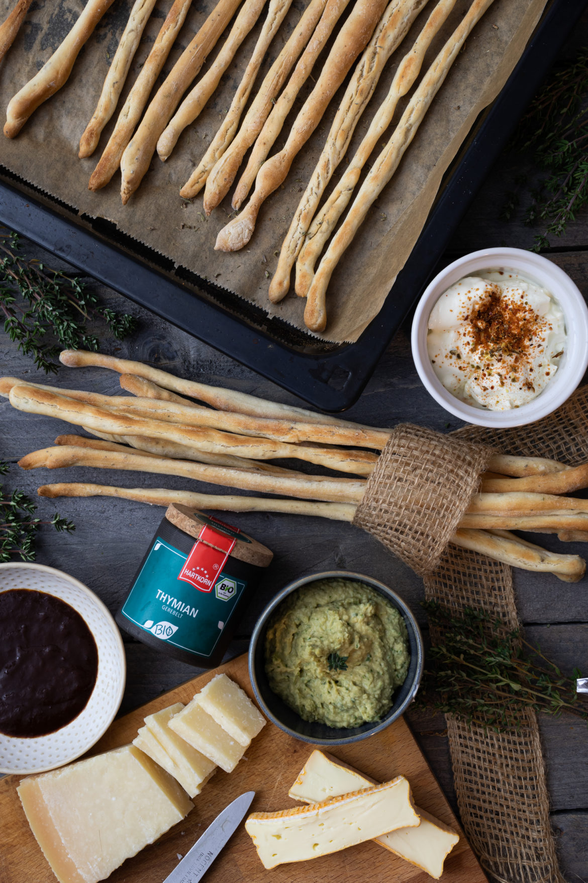 Knusprige Grissini auf Blech mit Dips und Käse auf Holzbrett, umgeben von frischen Kräutern.