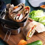 Wraps gefüllt mit Gemüse und Fleisch in einer Lunchbox auf einem Holzbrett, daneben eine Gurke und Salatblätter.