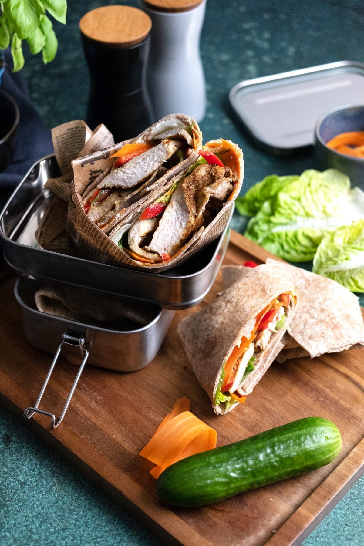 Wraps gefüllt mit Gemüse und Fleisch in einer Metall-Lunchbox auf einem Holzbrett, umgeben von Salat und Gemüse.