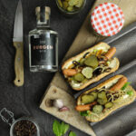 Zwei belegte Hotdogs mit Gurkenscheiben und Zwiebeln auf Holzbrett, daneben ein Messer, Ginflasche und Pfefferkörner.