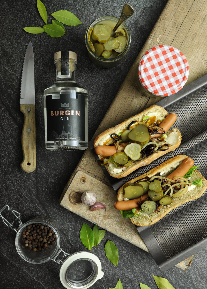 Zwei belegte Hotdogs mit Gurkenscheiben und Zwiebeln auf Holzbrett, daneben ein Messer, Ginflasche und Pfefferkörner.