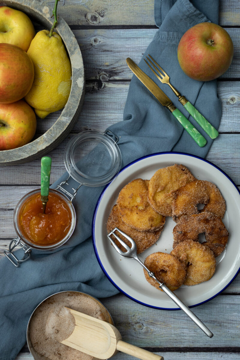 Gebratene Apfelringe auf Teller mit Gabel, Apfelmarmelade im Glas, Zimt-Zucker-Mischung, Äpfel und Zitrone in Holzschale.