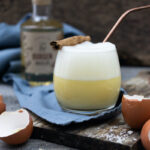 Ein Glas mit Schaum-Drink, Zimtstange und Metallstrohhalm, umgeben von Eierschalen und einer Flasche im Hintergrund.