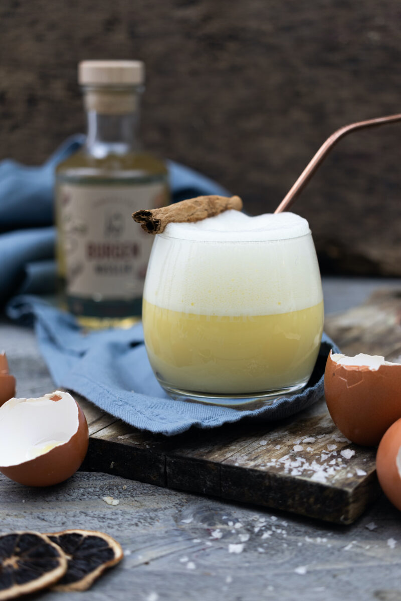 Ein Glas mit Schaum-Drink, Zimtstange und Metallstrohhalm, umgeben von Eierschalen und einer Flasche im Hintergrund.