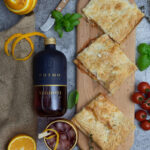 Flasche Negroni, Cocktail mit Eis, Orangen, Blätterteiggebäck, Tomaten und frische Kräuter auf grauem Untergrund.