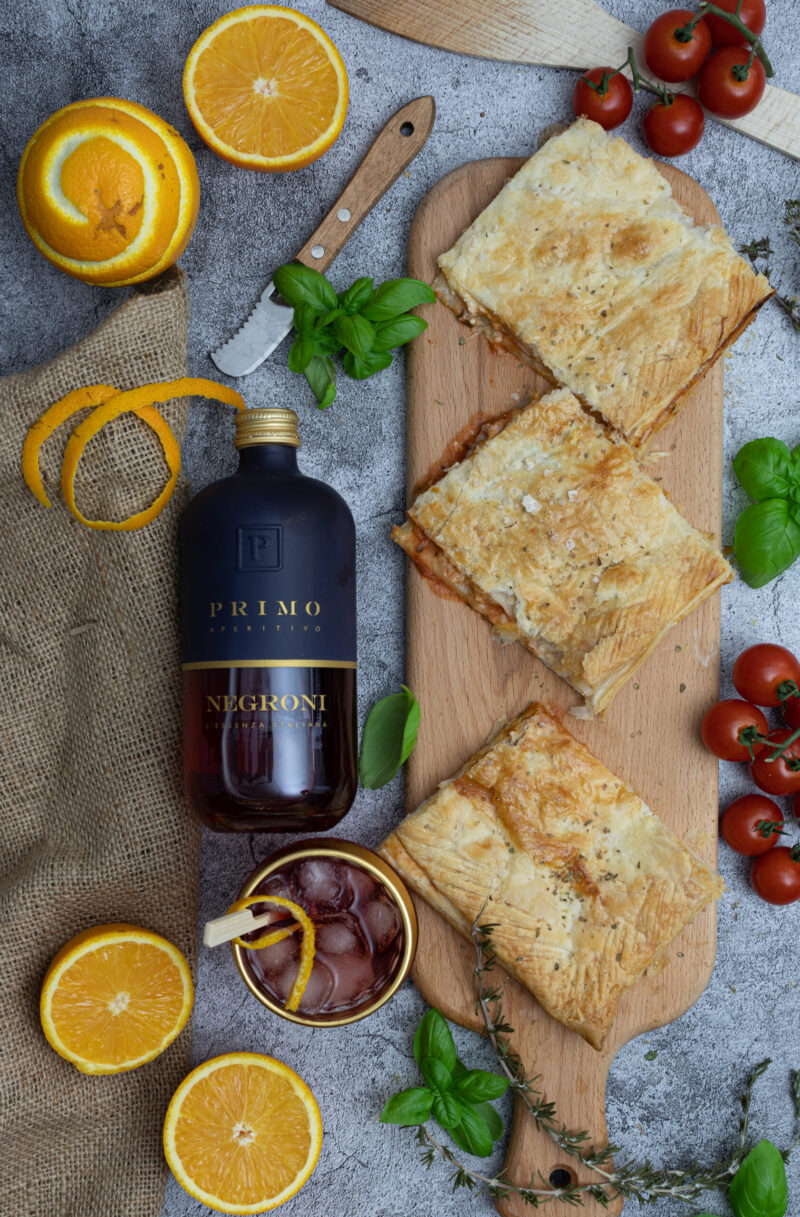 Flasche Negroni, Cocktail mit Eis, Orangen, Blätterteiggebäck, Tomaten und frische Kräuter auf grauem Untergrund.