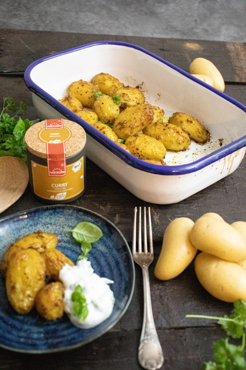 Gebackene Kartoffeln in einer Ofenform, daneben Currygewürz und ein Teller mit Kartoffeln und Joghurt-Dip.