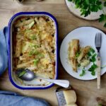 Überbackener Auflauf mit Frühlingszwiebeln in blauer Form, daneben ein Teller mit Portion, Käsereibe und Kräuter.