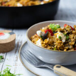 Eine Schüssel mit Gemüse-Reis und Feta. Daneben eine Gabel auf einem Teller. Im Hintergrund eine Pfanne.