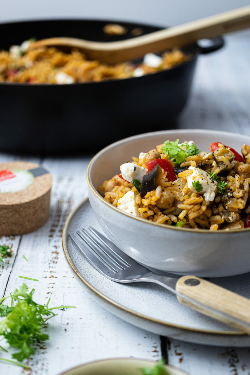 Eine Schüssel mit Gemüse-Reis und Feta. Daneben eine Gabel auf einem Teller. Im Hintergrund eine Pfanne.