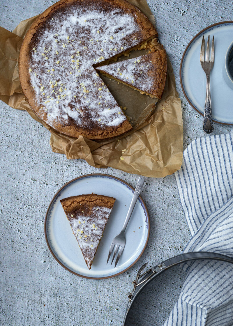 Kuchen mit Zuckerglasur und Zitronenabrieb, ein Stück herausgeschnitten. Teller mit Gabel daneben. Stoffserviette sichtbar.