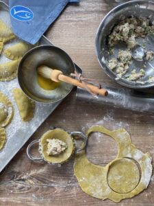 Teigkreise werden gefüllt und zu Pasteten geformt; Pinsel im geschmolzenen Fett, Schüssel mit Füllung und blauer Stoff.