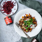 Teller mit geröstetem Blumenkohl, Karotten, Kichererbsen, Granatapfelkernen und Joghurt. Daneben Gewürze und Granatapfelkerne.