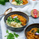 Drei Schalen mit Linsencurry und Reis, garniert mit frischen Kräutern und Frühlingszwiebeln, auf einem Tisch mit Besteck.