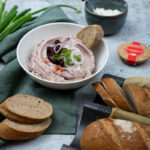 Schüssel mit Aufstrich, dekoriert mit Zwiebeln und Oliven, umgeben von Brotscheiben und Frühlingszwiebeln auf grauem Untergrund.