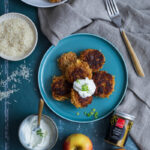 Ein blauer Teller mit Reisküchlein, garniert mit Joghurt, nebst Zutaten wie Äpfeln, Reis und Gewürz auf einem Tisch.