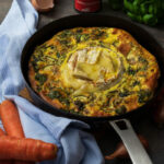 Frittata mit Spinat und Brie in schwarzer Pfanne, dekoriert mit Karotten, Eierschalen und Gewürzglas auf Holzuntergrund.