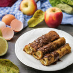 Drei goldbraune, schokoladenüberzogene Röllchen auf einem Teller, umgeben von Äpfeln und Zutaten auf einem Tisch.