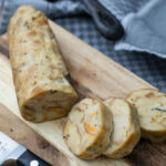 Gekochter Serviettenkloß in Scheiben geschnitten auf einem Holzbrett, daneben ein Messer.