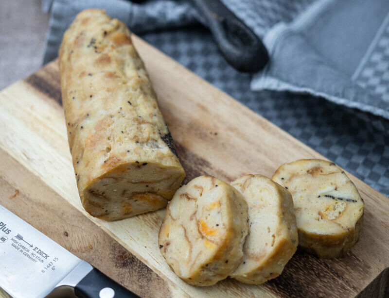 Gekochter Serviettenkloß in Scheiben geschnitten auf einem Holzbrett, daneben ein Messer.