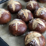 Ofenblech mit frisch gebackenen Laugenbrötchen, bestreut mit grobem Salz, auf Backpapier.