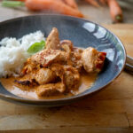 Dampfendes Hähnchengericht in Sahnesoße mit Reis auf einem Teller. Hintergrund: frische Karotten und Lauch.
