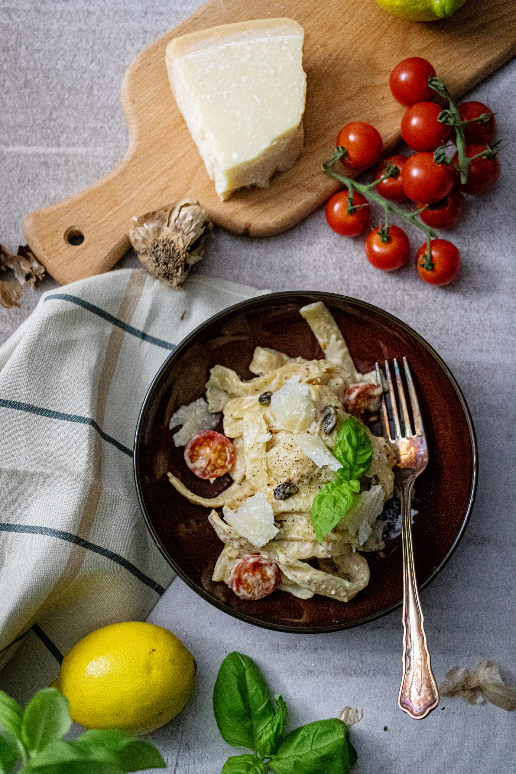 Pasta mit Sahnesoße, Kirschtomaten, Käse und Basilikum auf Teller. Daneben Käseblock, Tomaten, Zitrone und Tuch.