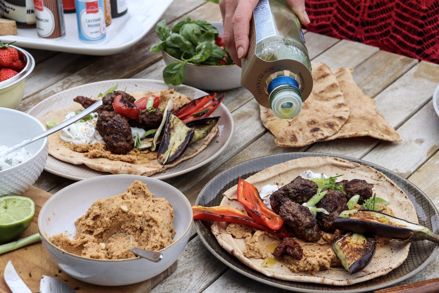 Hand gießt Öl über Fladenbrot mit gegrilltem Gemüse, Fleisch und Sauce. Zutaten und Schalen stehen auf Holztisch.