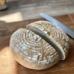 Ein halbiertes, rundes Bauernbrot mit Mehl bestäubt auf einem Holzbrett, daneben ein Brotmesser.