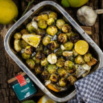 Gerösteter Rosenkohl mit Zitronenstücken und Knoblauch in einer Metallbackform auf Holztisch, umgeben von frischen Zutaten.