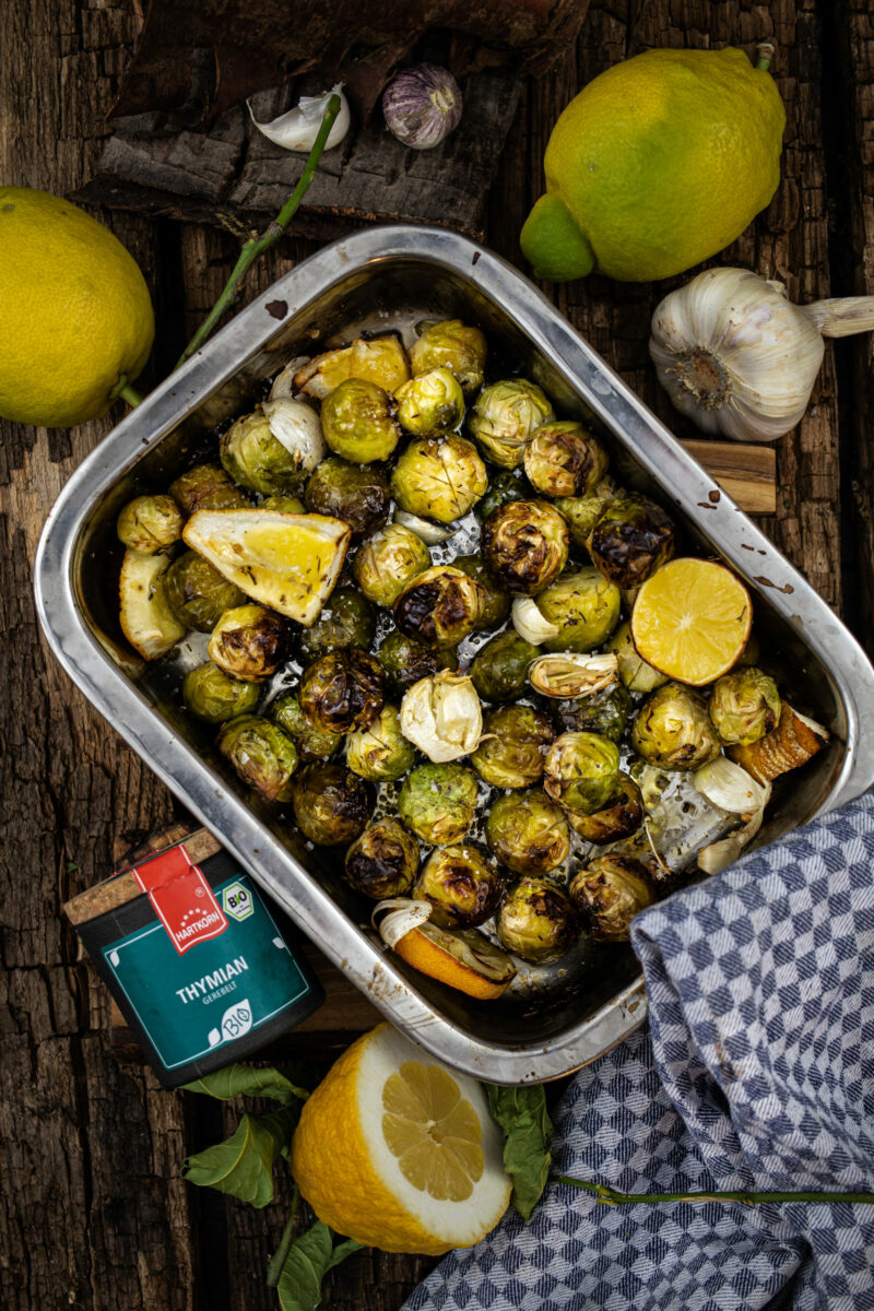 Gerösteter Rosenkohl mit Zitronenstücken und Knoblauch in einer Metallbackform auf Holztisch, umgeben von frischen Zutaten.