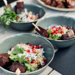 Schüsseln mit Reis, Granatapfelkernen, Kräutern und gegrillten Fleischspießen auf Holzuntergrund.