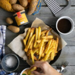 Pommes mit gewelltem Schnitt in Schale, Gewürz, Rohkartoffeln, Mühle, Soßen, Hand hält Pommes hält. Gabeln und Tuch daneben.