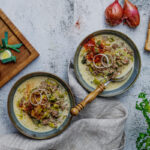 Zwei Teller mit Lauch-Hackfleisch-Suppe, neben Holzbrett mit Lauch, Knoblauch und Toast auf heller Unterlage.