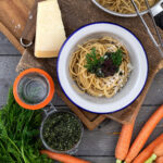 Schüssel Spaghetti mit Pesto, Kräutern und Käse, daneben Karotten, Parmesan und Glas auf Holzuntergrund.