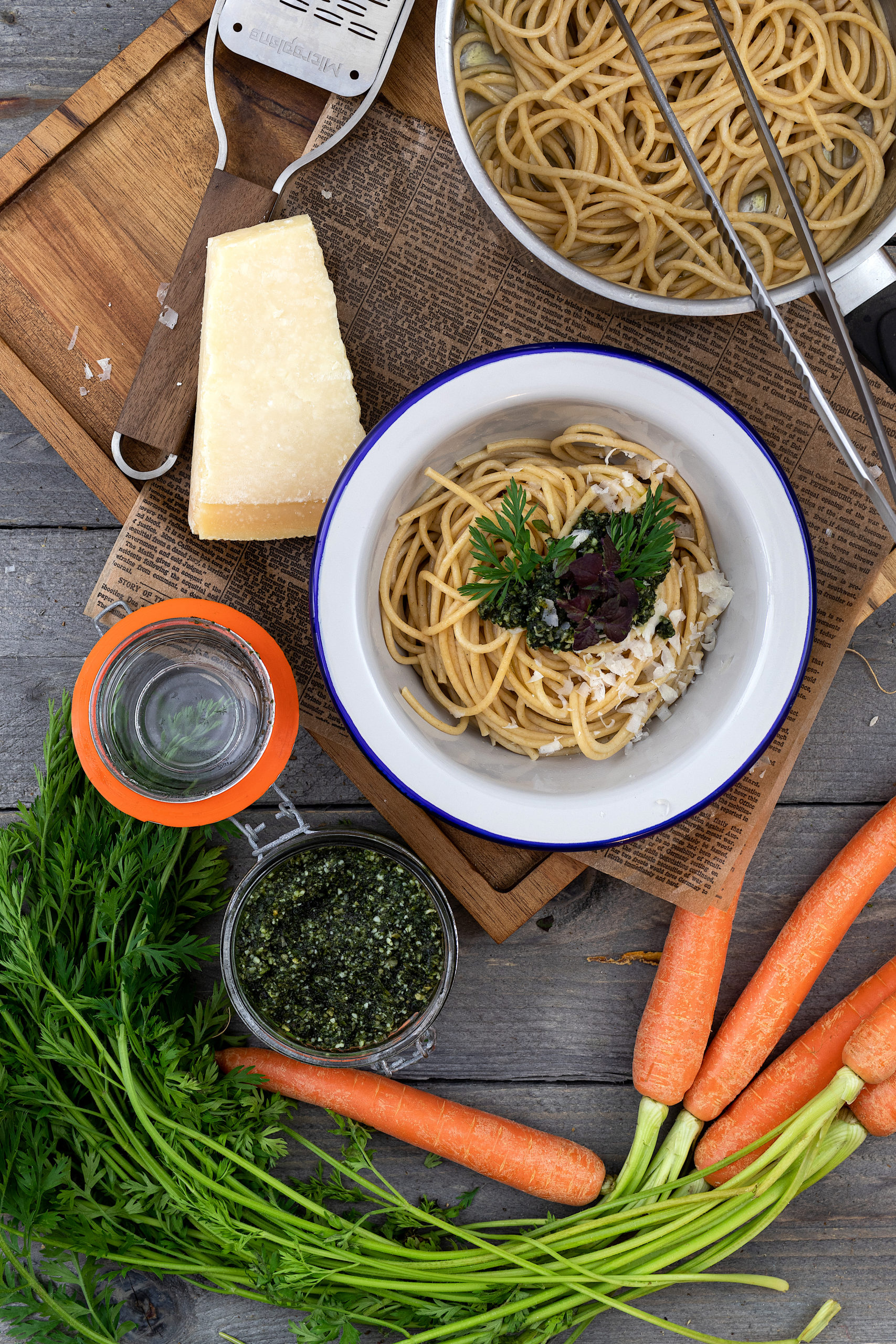 Schüssel Spaghetti mit Pesto, Kräutern und Käse, daneben Karotten, Parmesan und Glas auf Holzuntergrund.