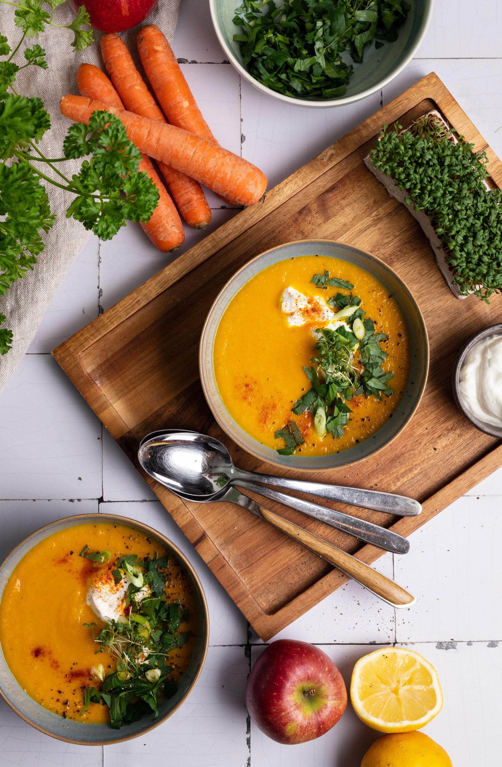 Zwei Schalen Karottensuppe mit Kräutern und Sahne auf Holzbrett, daneben Möhren, Apfel, Zitrone und frische Petersilie.