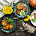 Drei Teller mit Kürbispuffern, frischen Kräutern und Creme. Ganze und geschnittene Kürbisse sind zu sehen.