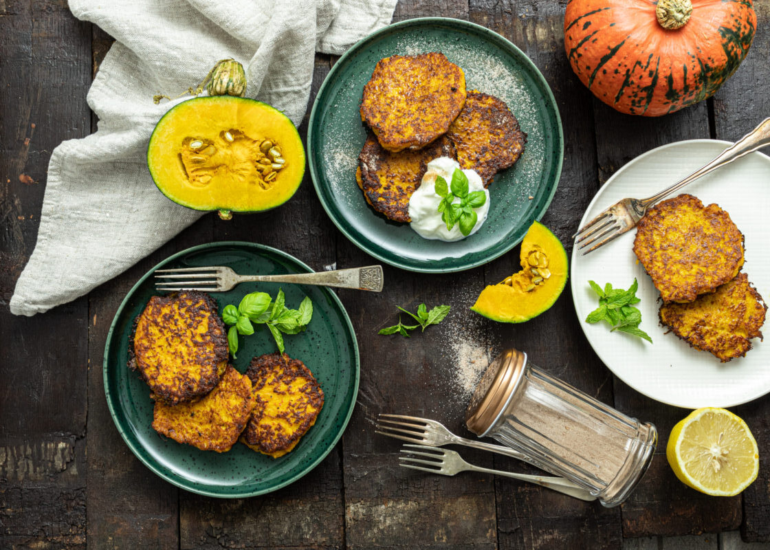 Drei Teller mit Kürbispuffern, frischen Kräutern und Creme. Ganze und geschnittene Kürbisse sind zu sehen.