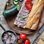Baguette mit Aufstrich, Gurkenscheiben und Tomaten auf Holzbrett. Daneben Schale mit Aufstrich, ganze Tomaten, Messer und Pfeffermühle.