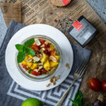 Glas mit Mango-Salat, Minze und Nüssen auf Teller, daneben Zitrone, Pfeffer und Zeitungsausschnitt auf grauem Tuch.