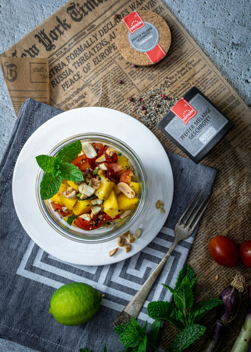 Glas mit Mango-Salat, Minze und Nüssen auf Teller, daneben Zitrone, Pfeffer und Zeitungsausschnitt auf grauem Tuch.