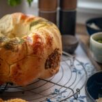 Geflochtenes Brot mit Gewürzkruste auf einem Gitter, umgeben von Schüsseln und Kräutertopf im Hintergrund.